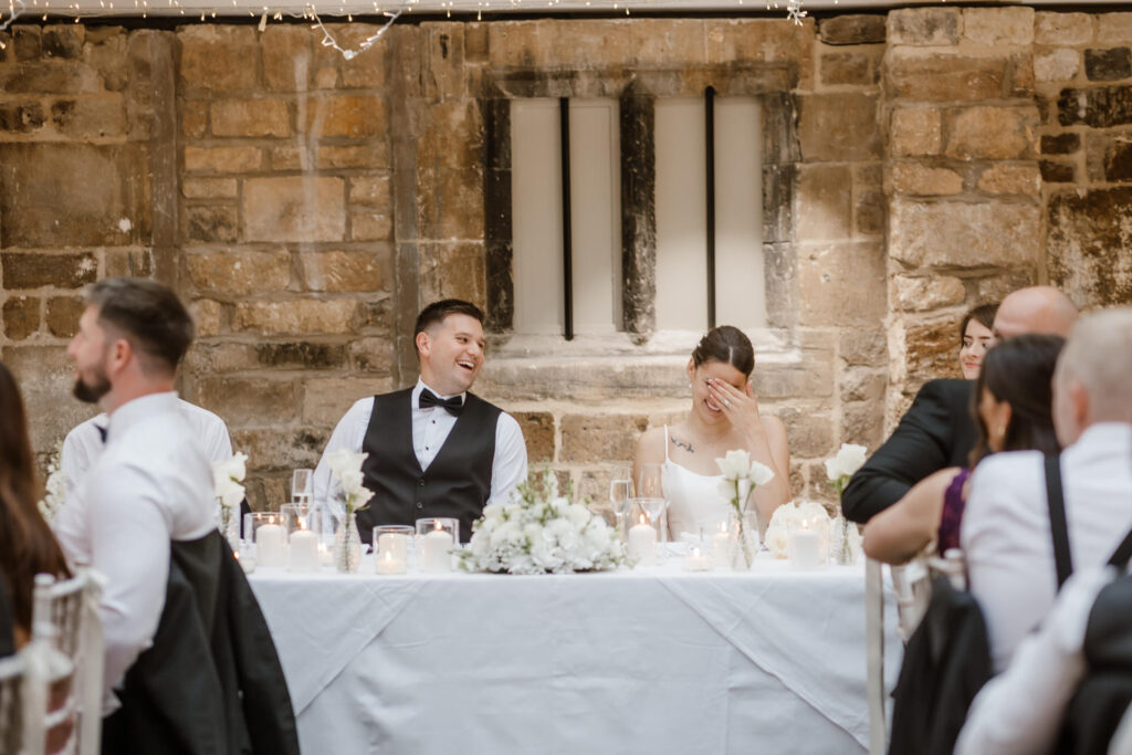 Wedding reception with guests seated at a head table. The couple, dressed formally, are laughing and smiling. The setting includes stone walls and fairy lights.