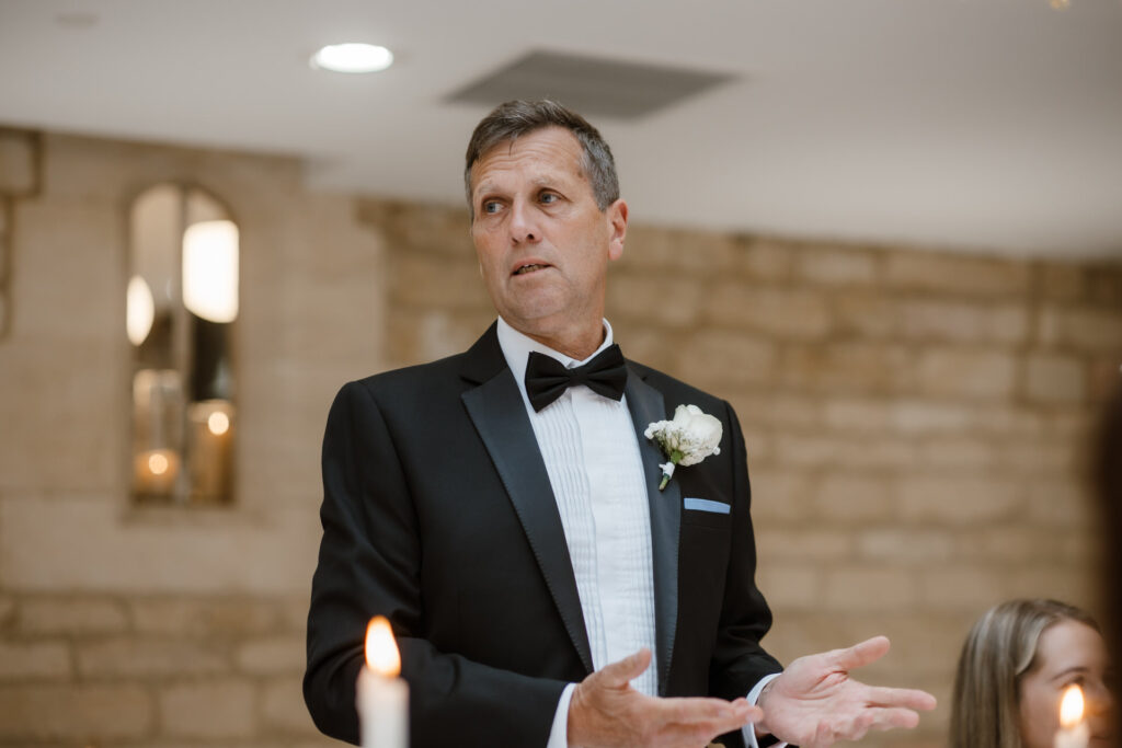 A man in a tuxedo and bow tie gives a speech in a stone-walled room, with a lit candle in the foreground.