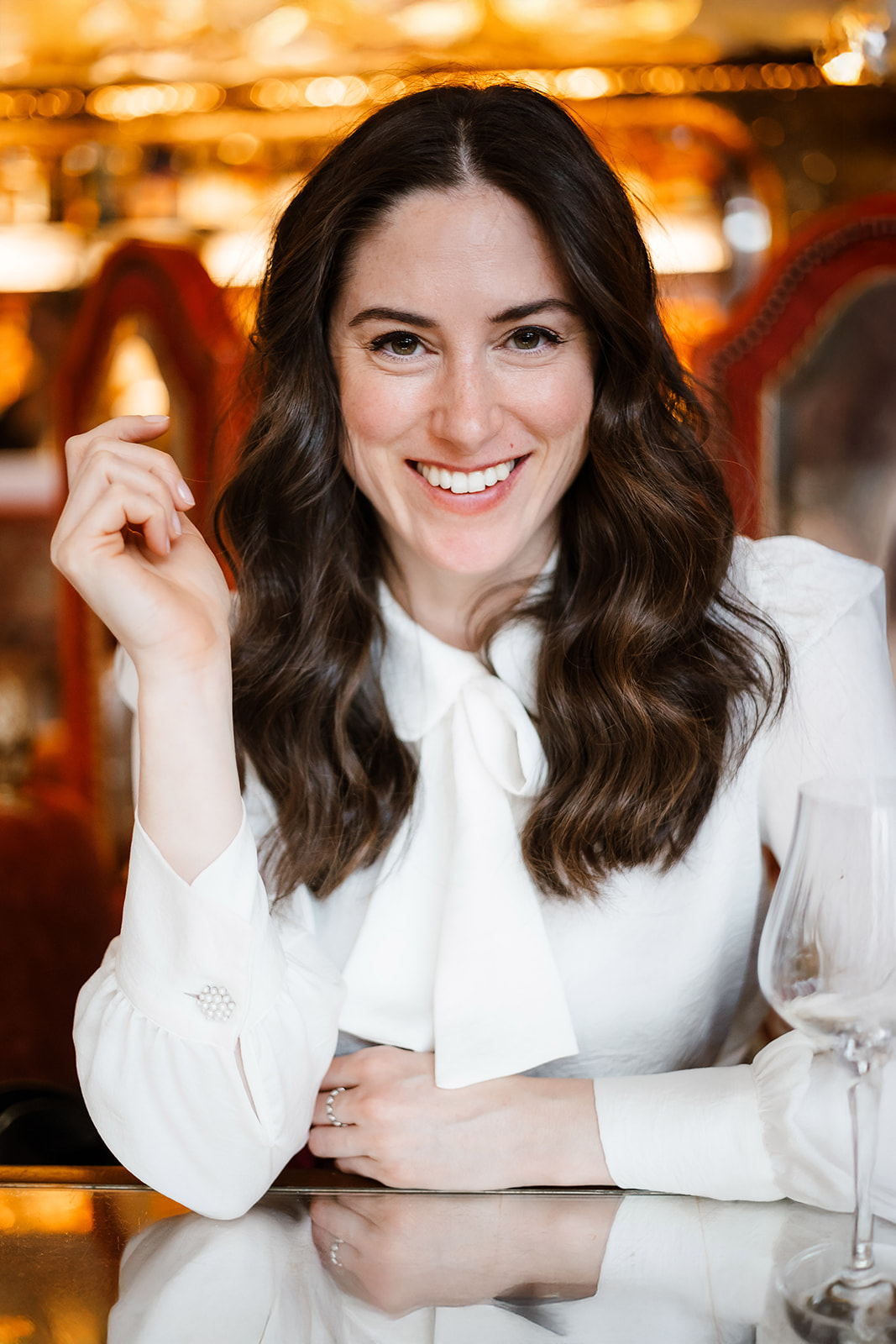 Birmingham Wedding Photographer Eva Murko Photography: A woman with long brown hair and a white blouse sits at a table with empty glassware, smiling at the camera in a warmly lit setting.