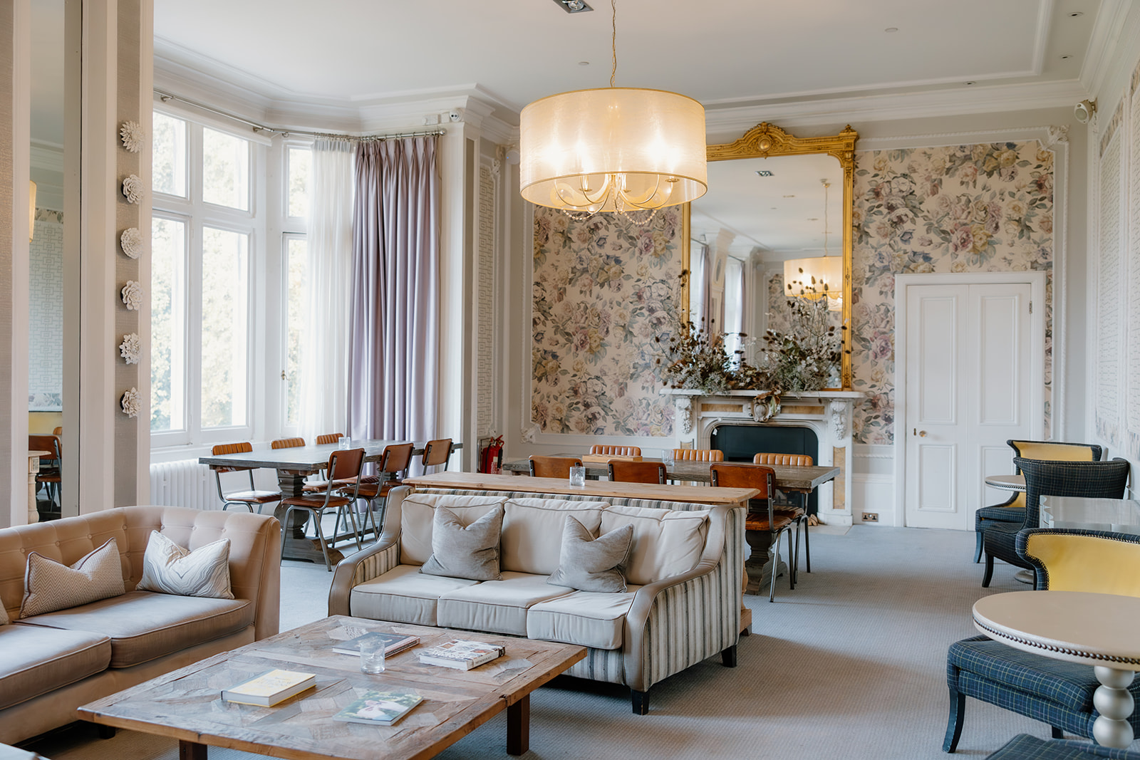 Elegant lounge called Tasting Room with sofas, armchairs, wooden tables, a large floral-patterned mirror above a fireplace, and a modern chandelier. Large windows let in natural light at Hampton Manor Solihull.