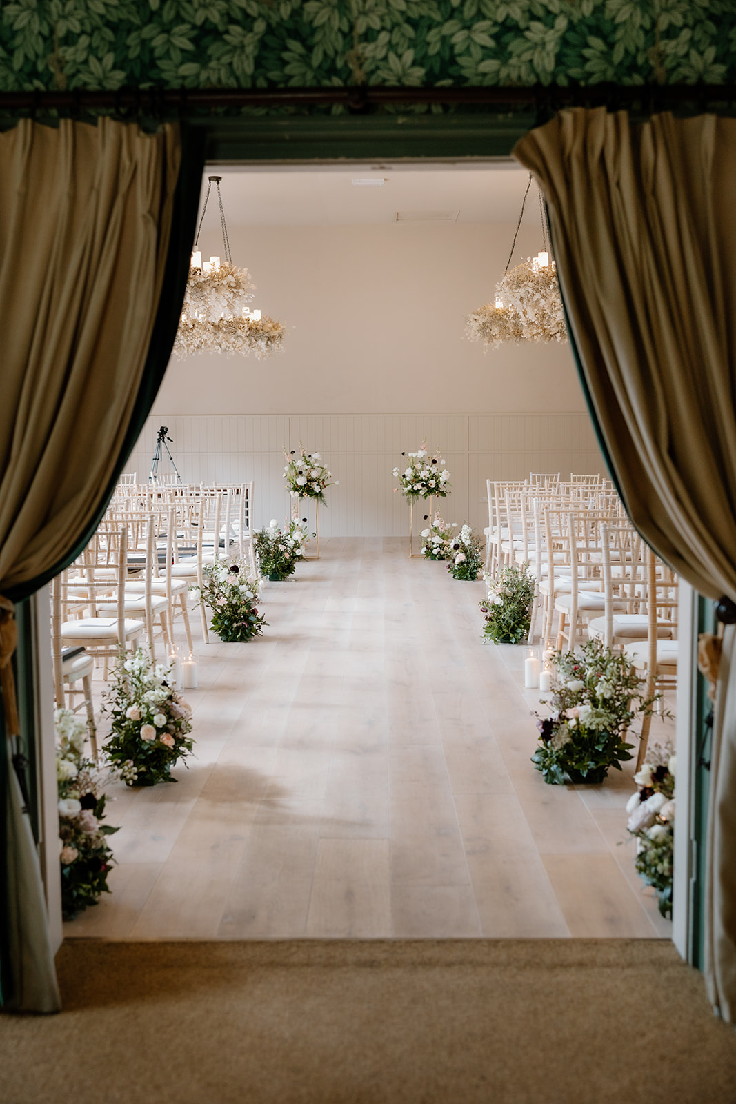 An elegant indoor wedding ceremony setup with rows of white chairs, floral arrangements lining the aisle, and chandeliers overhead, viewed through open curtains at The Birches.