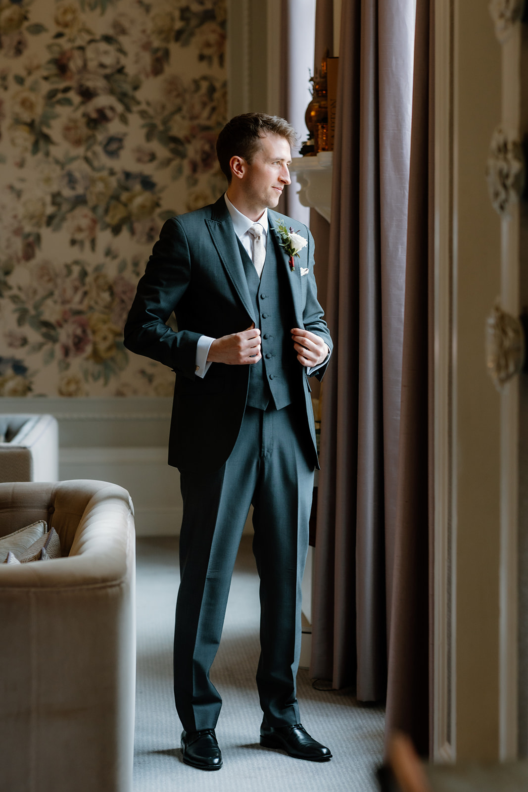 The groom in a suit stands by a window, adjusting his jacket, in a room with floral wallpaper and neutral-coloured furniture.