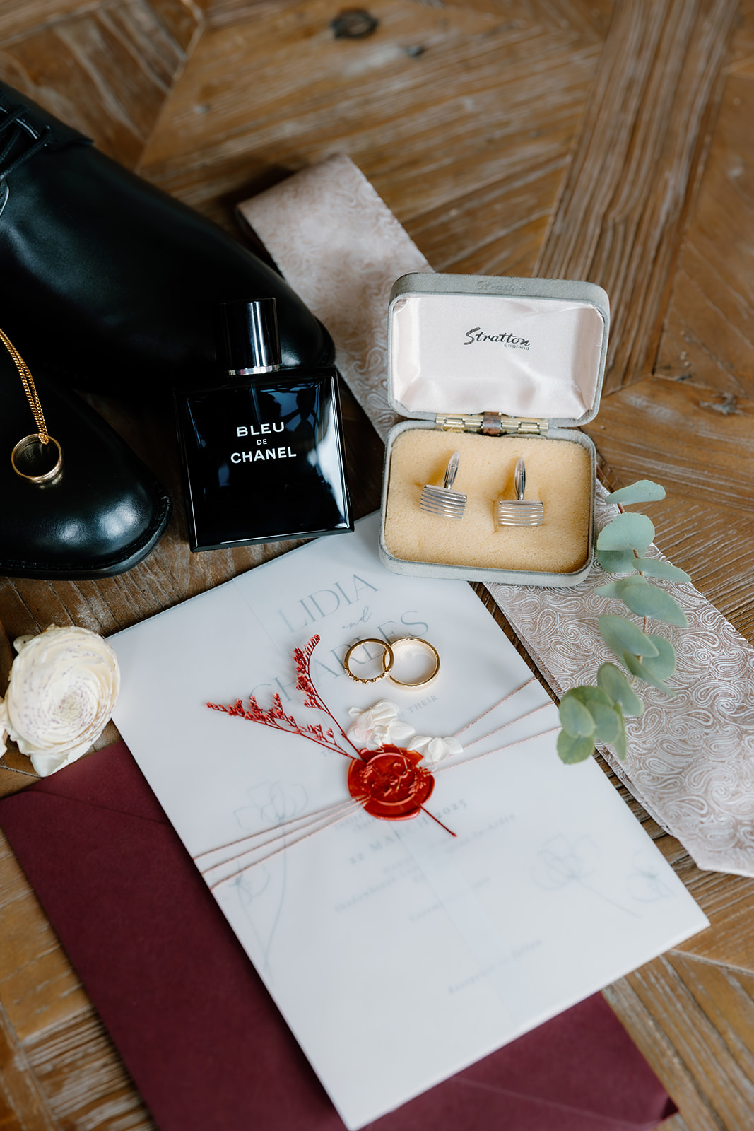 A flat lay of wedding accessories including black shoes, Bleu de Chanel cologne, cufflinks in a box, two rings, a wax-sealed envelope, a boutonnière, and a lace ribbon on a wooden surface.