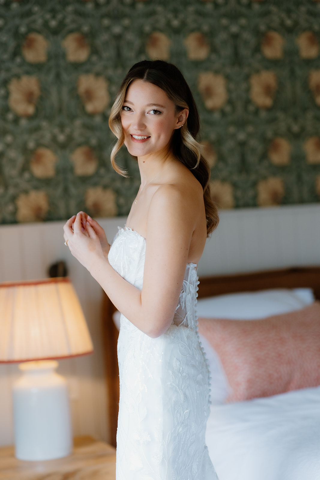 A woman in a strapless white lace wedding dress stands indoors, smiling, with a bed and lamp in the background.