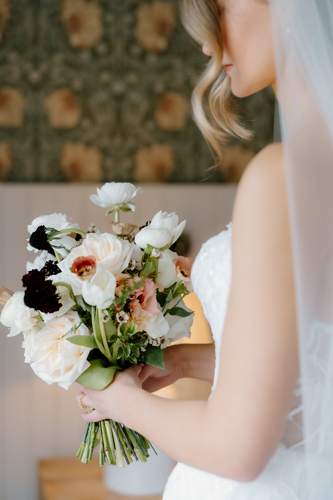A bride in a white dress holds a bouquet of white, pink, and dark flowers, with her face partially turned away from the camera.