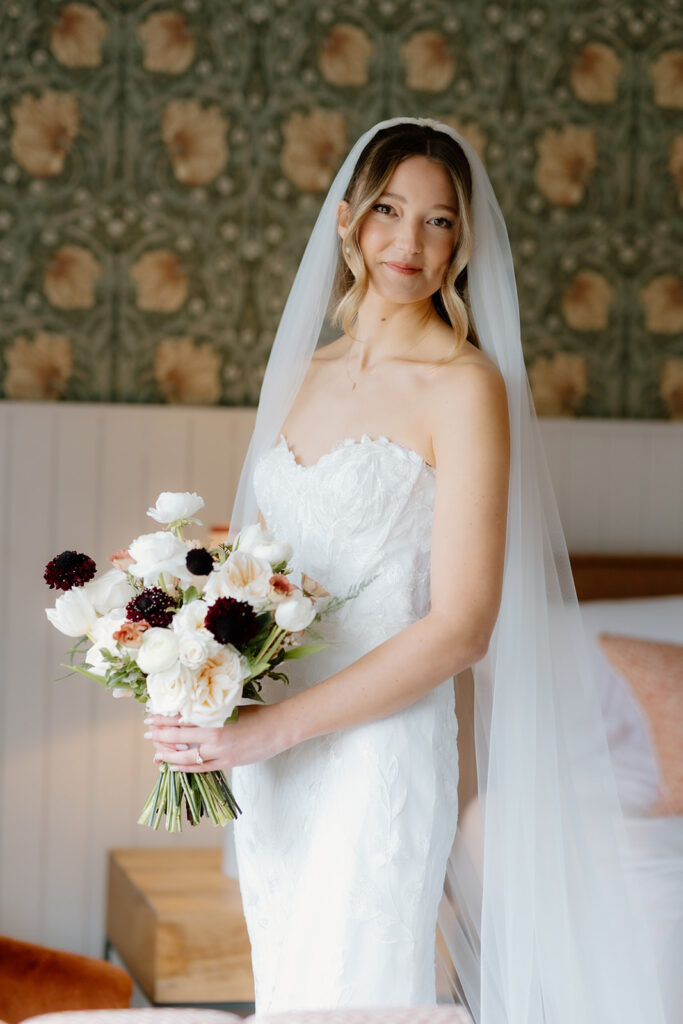 A bride in a strapless white wedding gown and veil stands indoors, holding a bouquet of white and dark flowers, with floral wallpaper in the background.