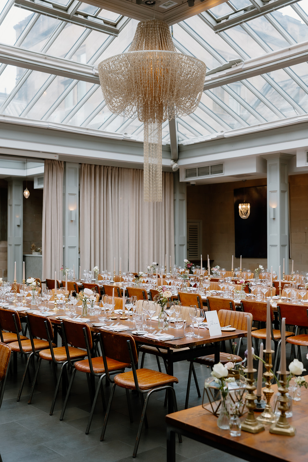 A formal dining room with long tables set for an event, featuring glassware, candles, floral centerpieces, wooden chairs, and a large chandelier under a glass ceiling.