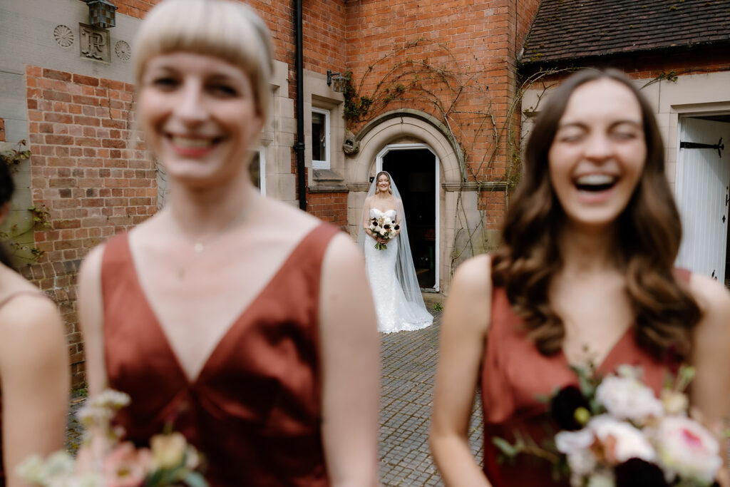A bride in a white gown stands in a doorway holding a bouquet while two blurred bridesmaids in rust-colored dresses smile and laugh in the foreground.