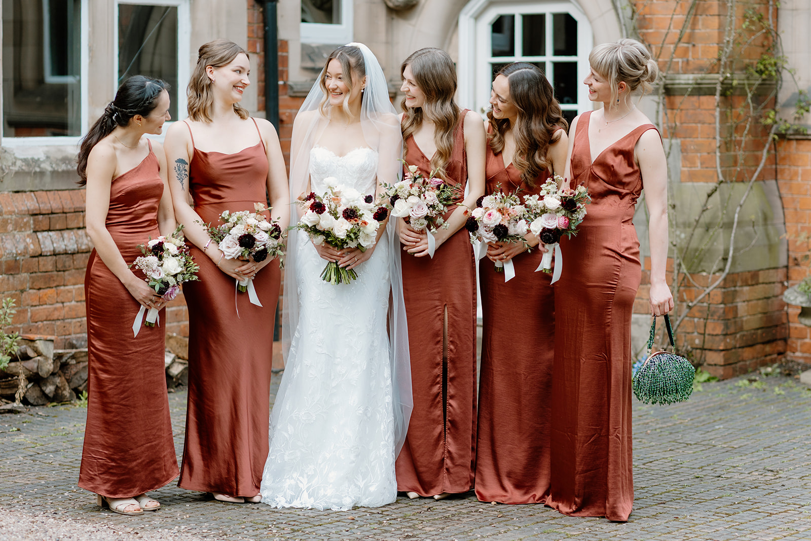 Lidia & Charles A bride in a white wedding dress stands outside with five bridesmaids wearing matching rust-colored dresses, all holding bouquets and smiling at each other.