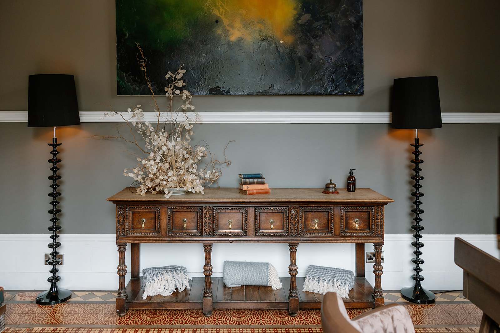 A wooden console table with drawers is topped with books, a vase of white branches, and a bottle, flanked by black floor lamps beneath a dark abstract painting.