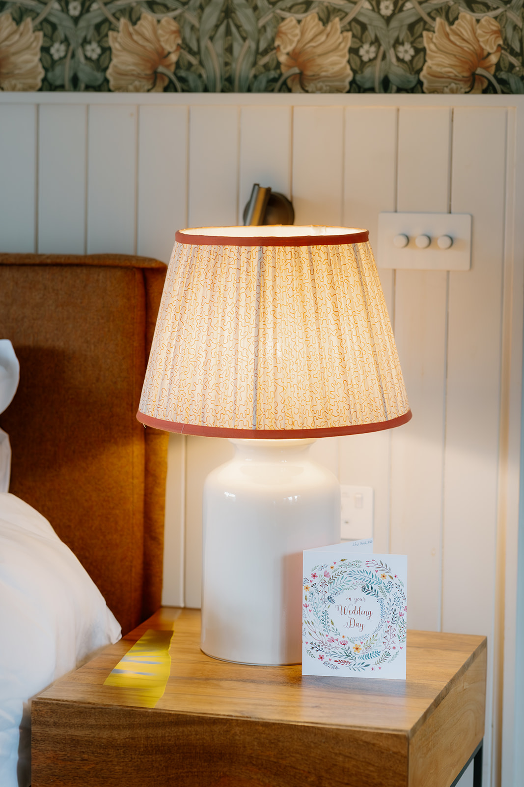 A lit table lamp with a patterned shade sits on a wooden nightstand next to a greeting card and a yellow ribbon in a bedroom setting.