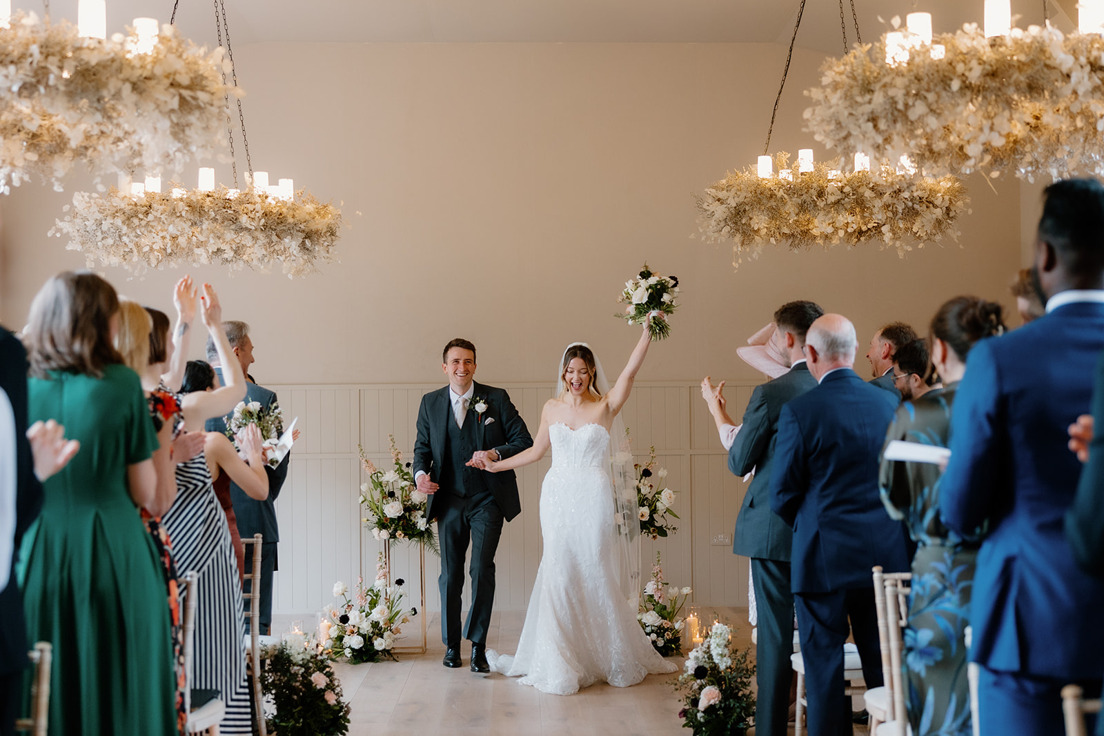 A bride and groom walk down the aisle together as guests stand and applaud in a decorated indoor wedding ceremony room The Birches.