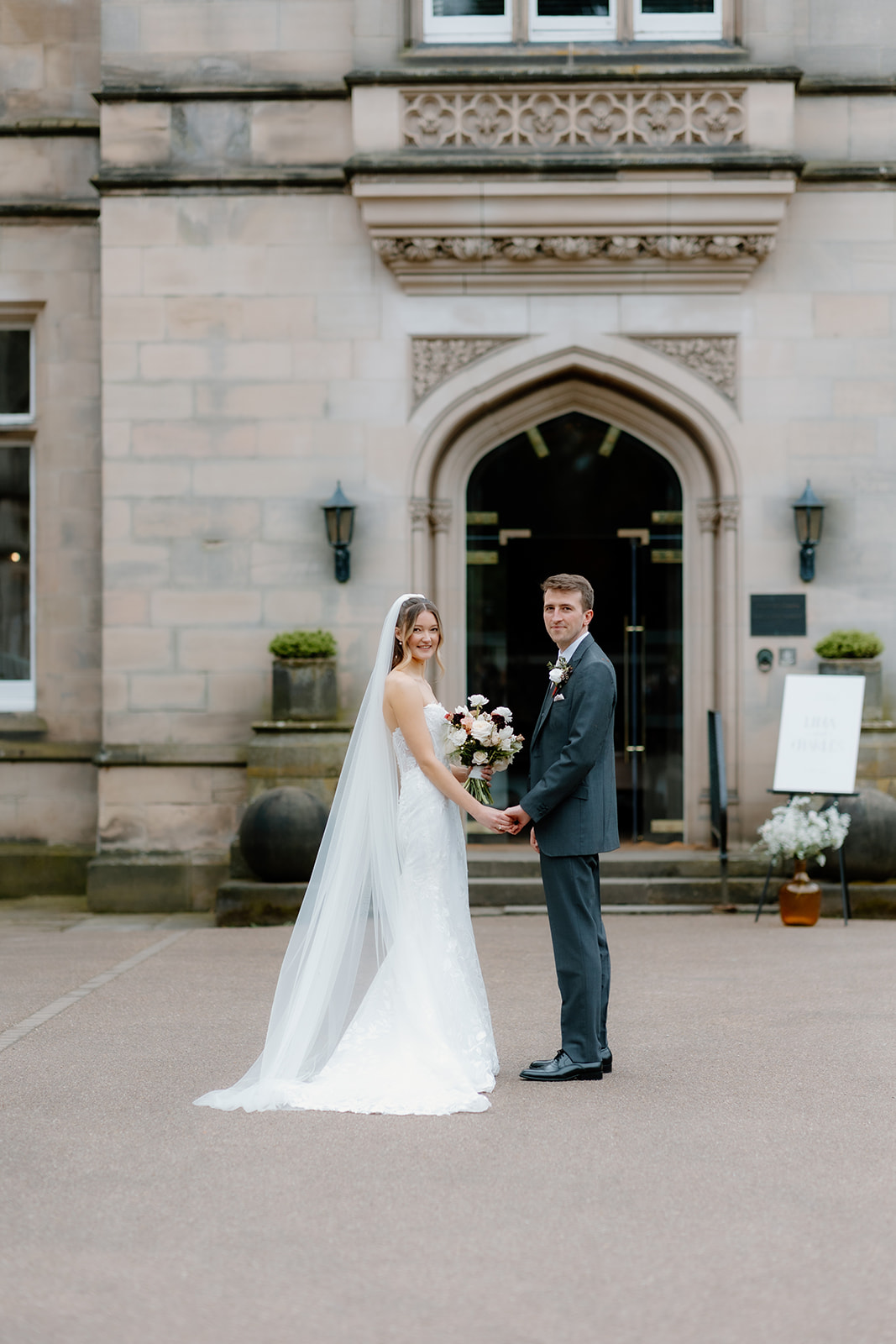 Lidia & Charles A bride and groom stand hand in hand in front of a stone building entrance, both looking at the camera. The bride wears a white dress and veil; the groom wears a dark suit.