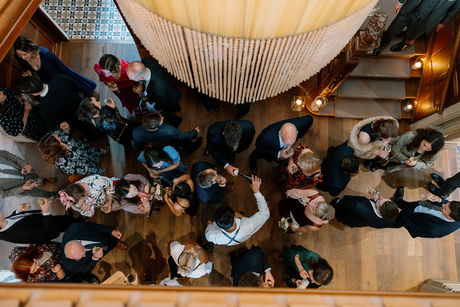 Overhead view of a group of people dressed formally, socializing and holding drinks in a wooden-floored room near a staircase and chandelier.