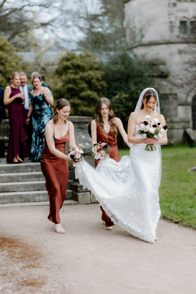 A bride in a white dress walks outdoors with two bridesmaids in rust-colored dresses holding bouquets and helping carry her train, with guests standing on stone steps in the background.