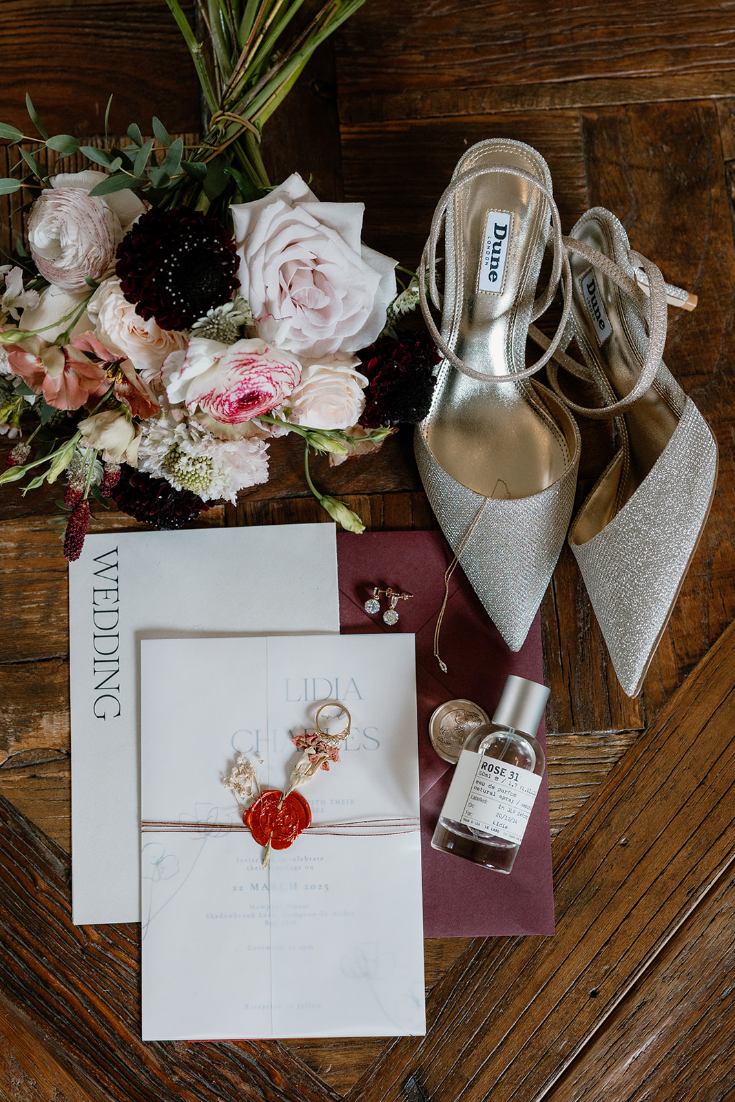 Flat lay of wedding essentials on a wooden surface, including silver shoes, flowers, invitation, envelope, perfume, jewelry, rings, and a wax-sealed letter.