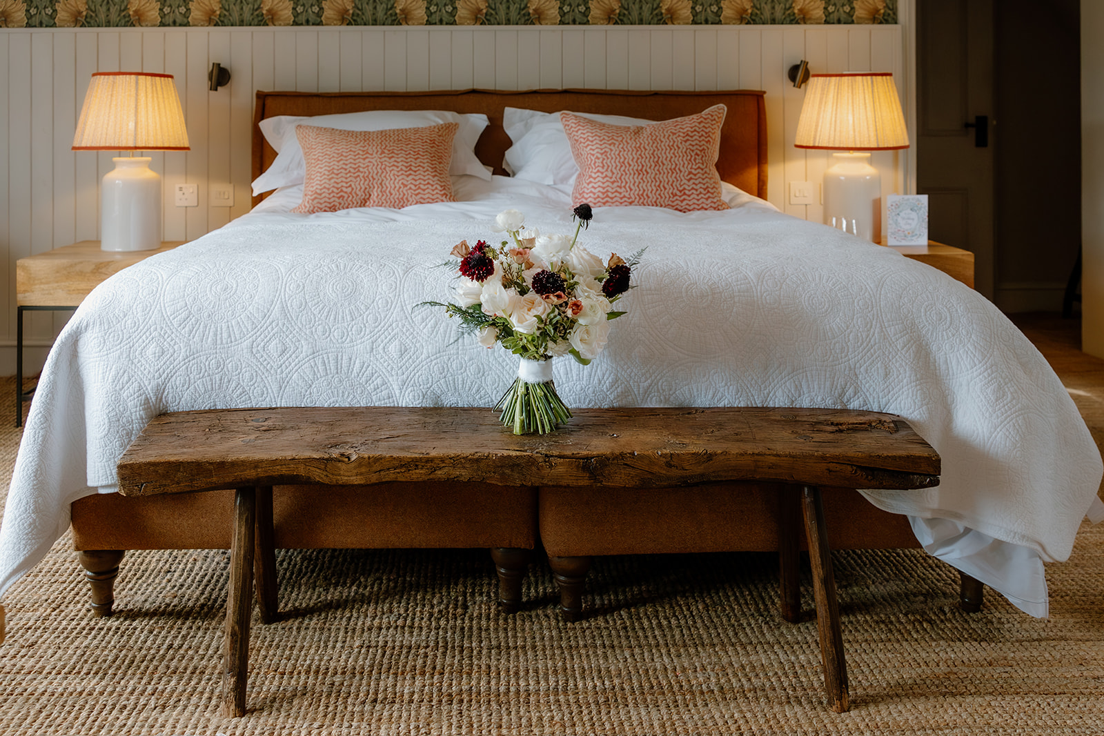 A neatly made bed with white bedding, two lamps on bedside tables, two decorative pillows, a floral arrangement on a rustic wooden bench, and a textured rug on the floor at Manor Cottage.
