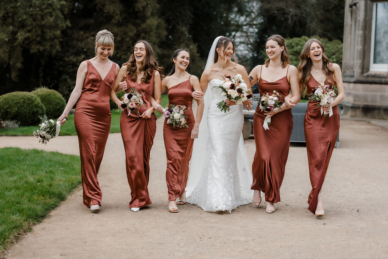 A bride in a white dress walks outside with five bridesmaids wearing matching rust-colored dresses, all holding bouquets and smiling.