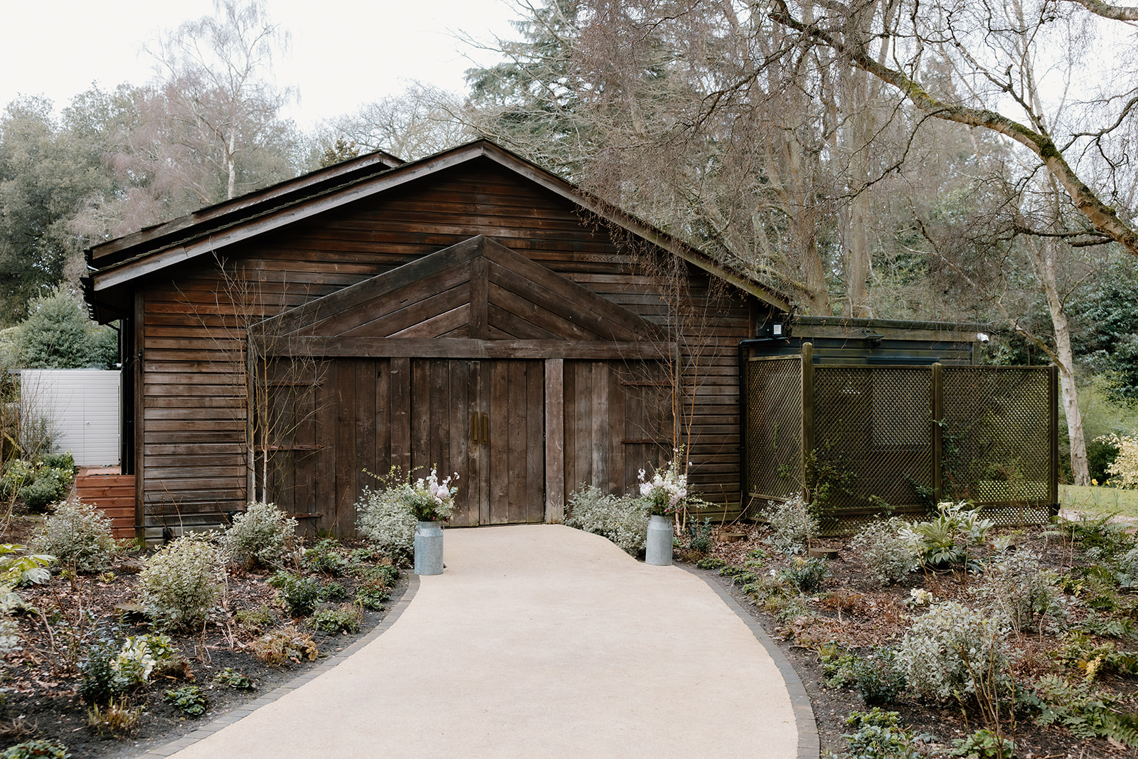 A wooden building The Birches with double doors is surrounded by landscaped plants and trees, with a paved pathway leading to the entrance.