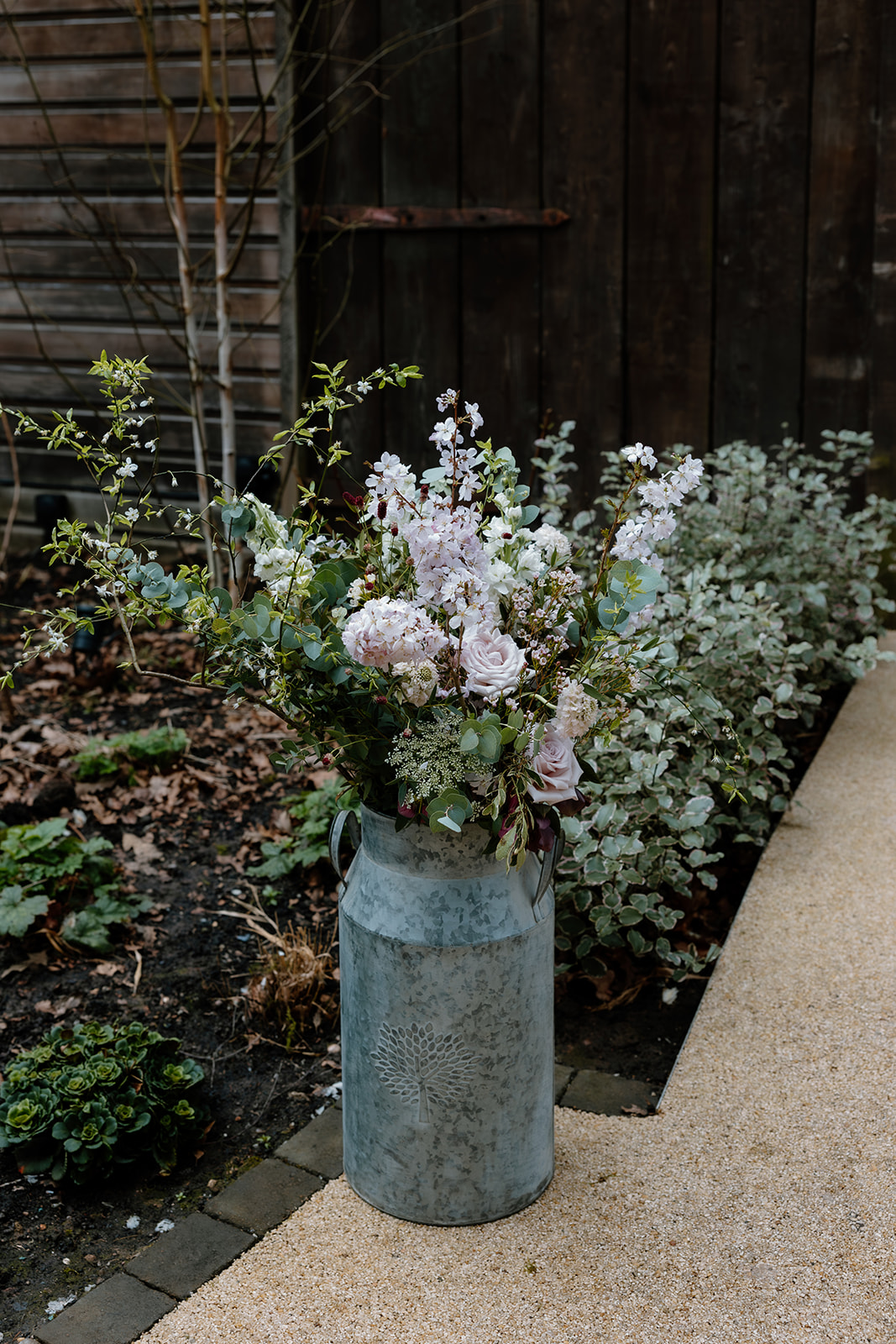 A large galvanized metal milk can filled with assorted flowers and greenery stands on a garden path next to a planted area and wooden fence.