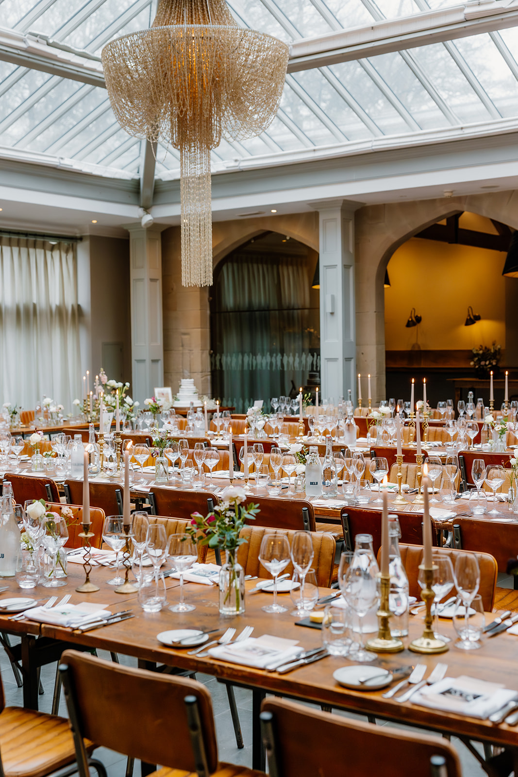 The elegant banquet hall at Hampton Manor wedding venue has long wooden tables set with glassware, plates, candles, and floral centrepieces under a large chandelier and glass ceiling.