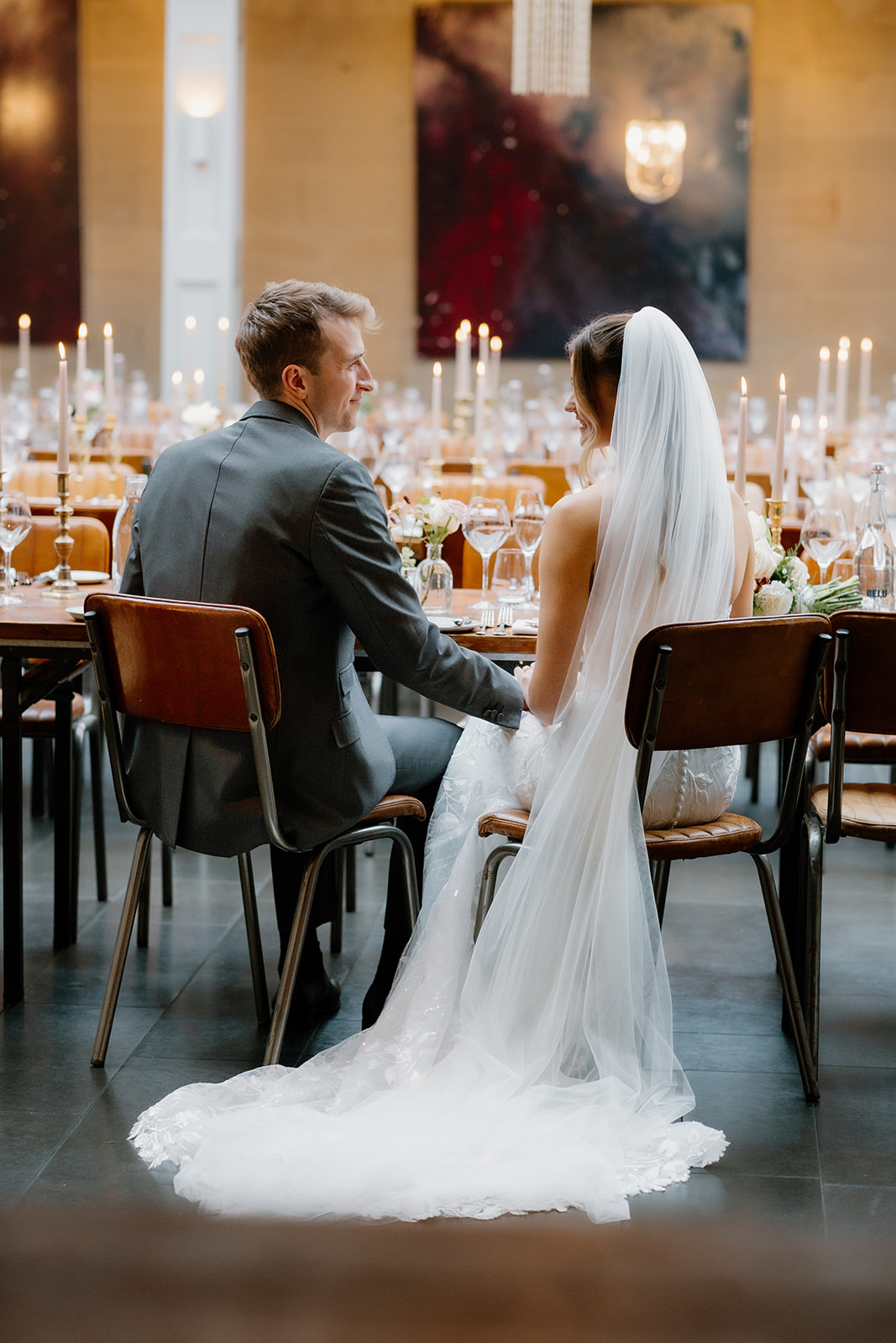 Lidia & Charles A bride and groom sit side by side at a decorated table, holding hands and facing each other, with candles and place settings visible around them.