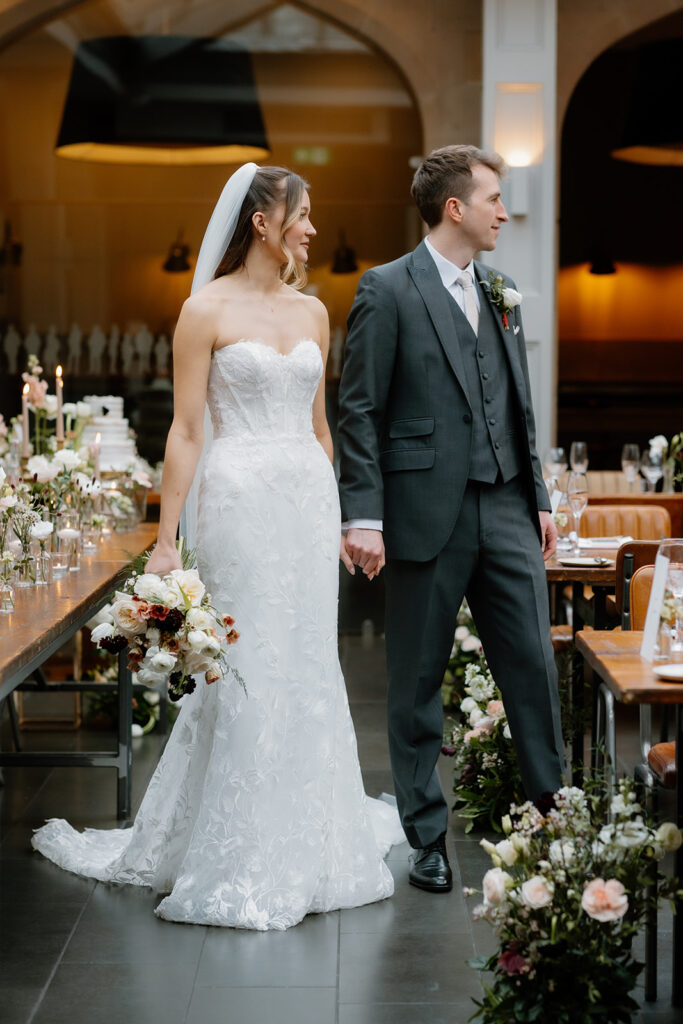 A bride in a white strapless gown and a groom in a gray suit hold hands and stand in a decorated wedding venue with floral arrangements and set dining tables.