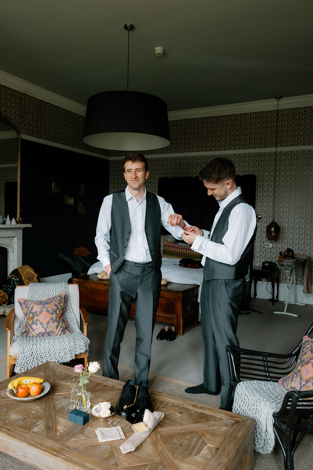 Two men in dress shirts and vests stand in a well-furnished bedroom, one adjusting the other's cufflink. Clothes, shoes, and items are laid out on a wooden coffee table in the foreground.