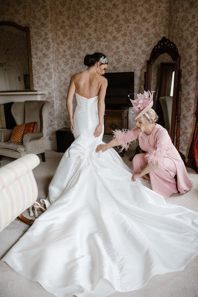 A woman in a strapless wedding dress stands as another woman in a pink outfit adjusts the train of the gown in a decorated room.