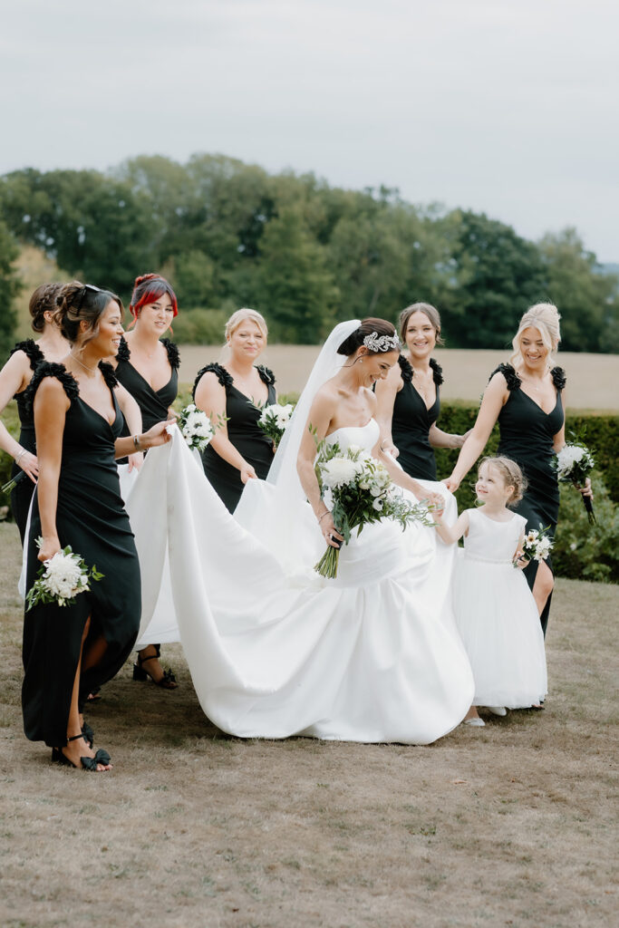 A bride in a white gown walks outdoors with her bridesmaids in black dresses and a young flower girl in a white dress.