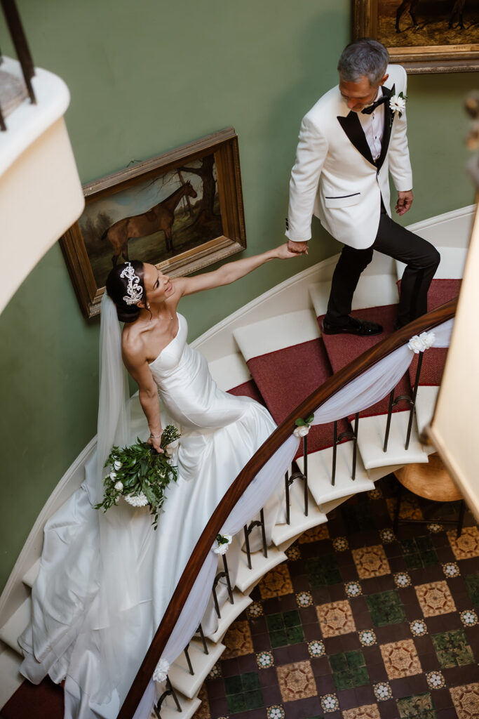 A bride in a white gown and veil holds a bouquet while ascending a spiral staircase, reaching for the groom in a white tuxedo jacket and black pants.