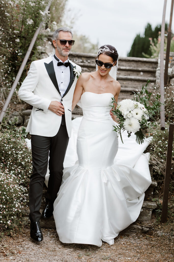 A bride in a strapless white gown and a man in a white tuxedo jacket walk down outdoor stone steps, both wearing sunglasses. The bride holds a bouquet of white flowers.