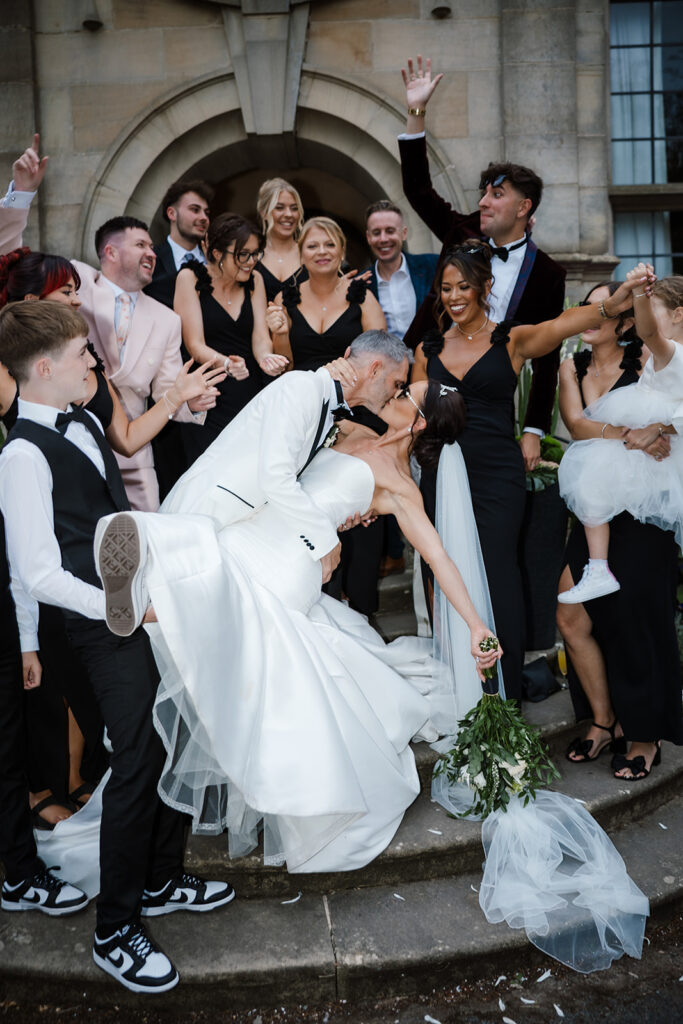 A newlywed couple kisses as the groom dips the bride, surrounded by cheering wedding guests on stone steps outside a building.