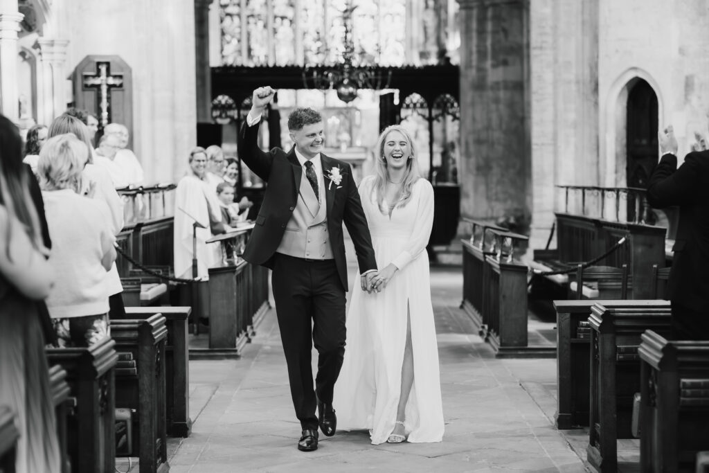 A newlywed couple walks down the aisle in a church, holding hands and smiling, as guests look on and celebrate.
