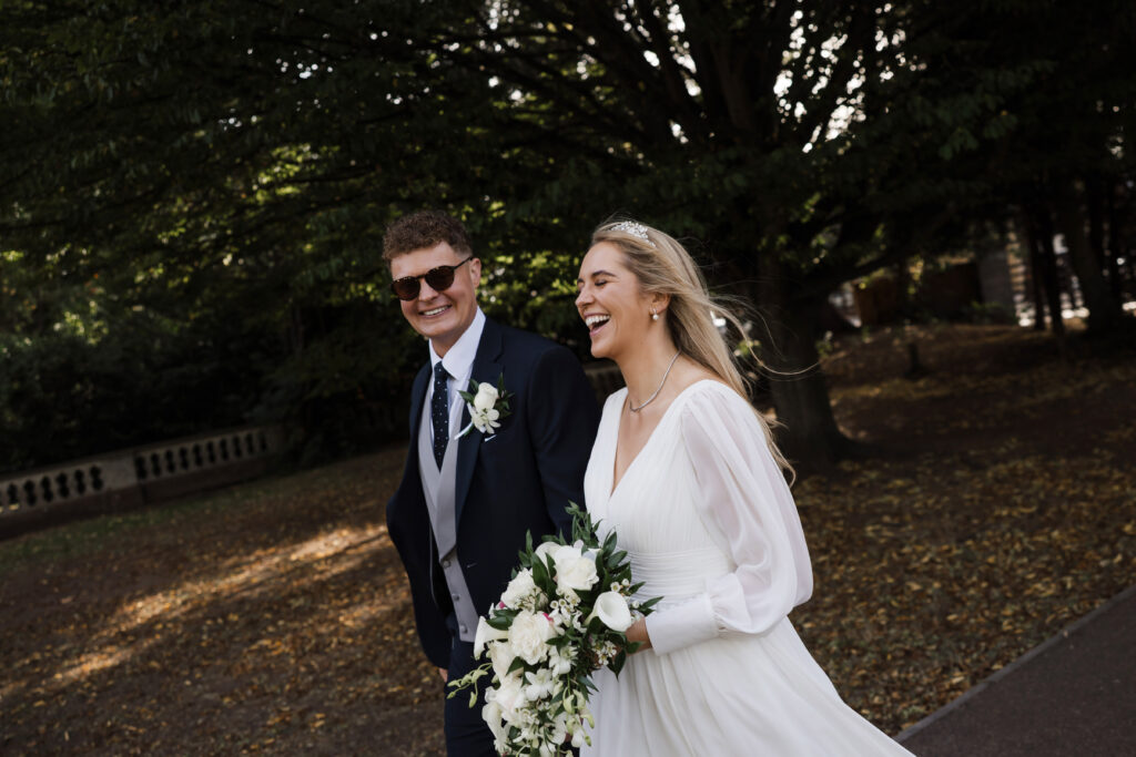 A bride in a white dress and a groom in a suit walk outdoors together, smiling and holding a bouquet, with trees and fallen leaves in the background.