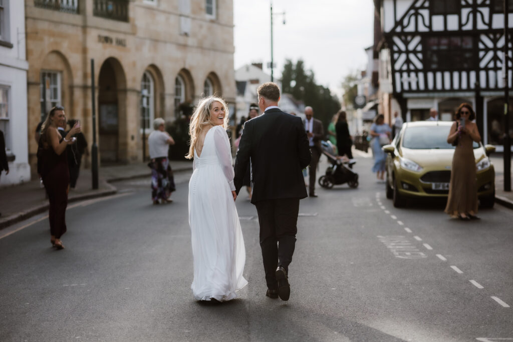 A bride and groom walk hand in hand down a street, with onlookers and parked cars around them, as the bride looks back and smiles.