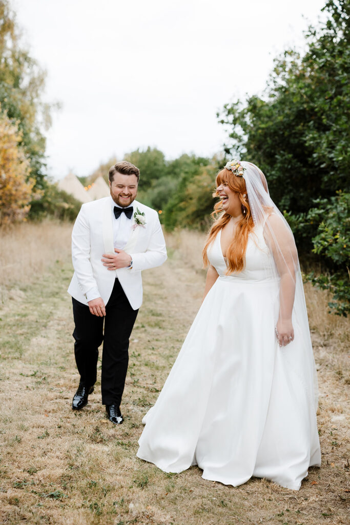 A bride and groom walk outdoors on a grassy path, dressed in formal wedding attire, both smiling and laughing together.