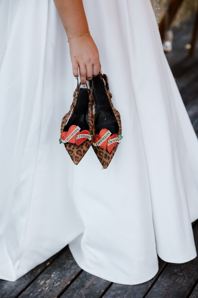 A person in a white dress holds a pair of leopard print shoes with embroidered red heart patches and the word "STRANGE" on them.