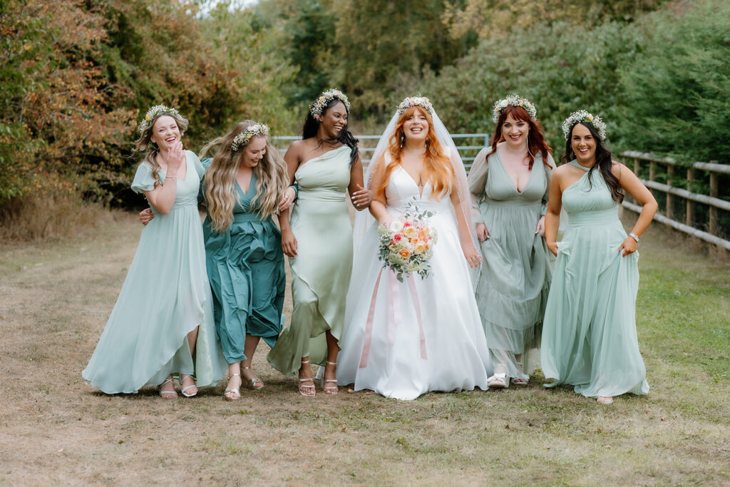 Six women in light pastel dresses, including a bride in a white gown holding a bouquet, walk together outdoors on a grassy path, smiling and laughing.