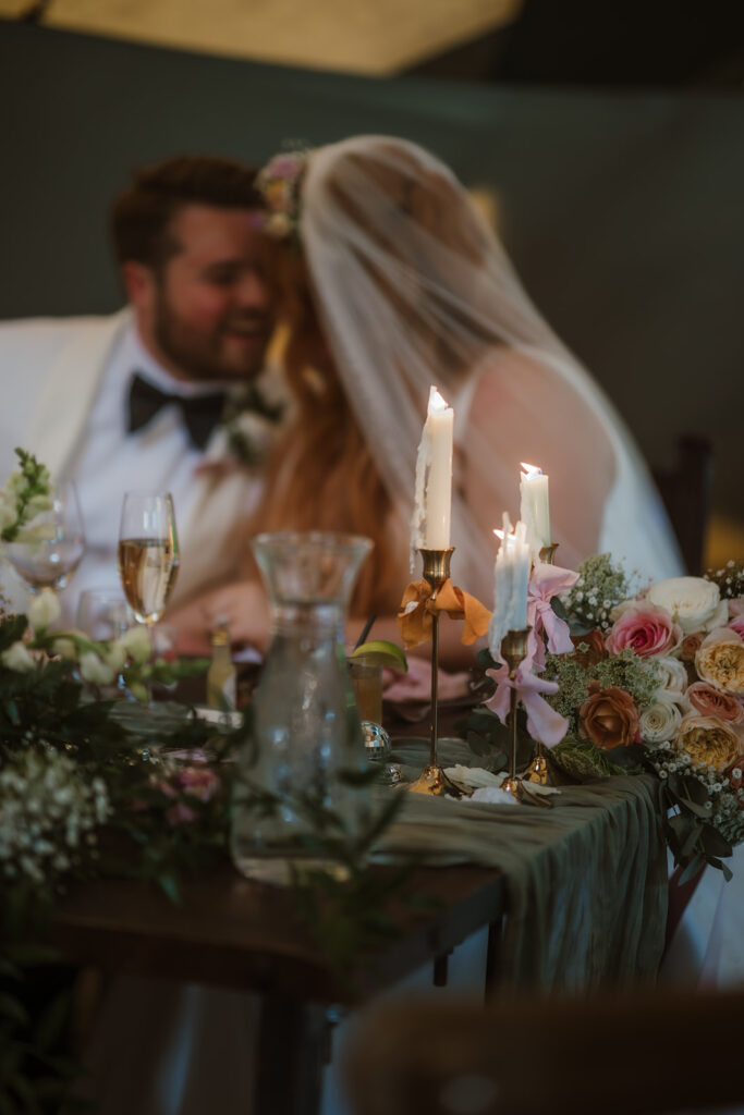 Bride and groom sit closely together at a decorated table with flowers, candles, and glasses, with their faces partially out of focus.