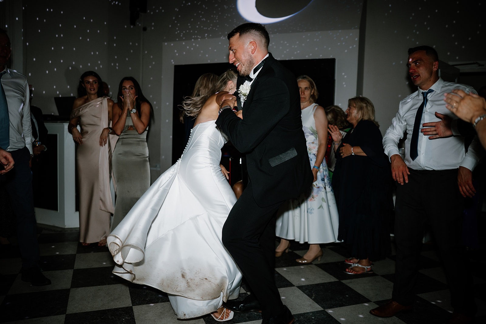A bride and groom dance energetically on a chequered floor, surrounded by guests in formal attire under dim lighting with light spots on the walls.