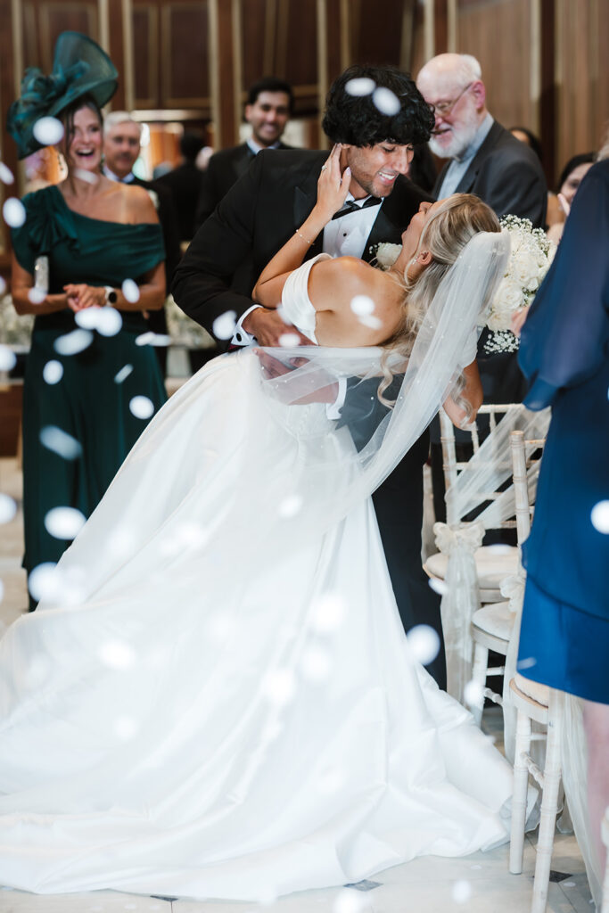A groom dips and kisses the bride at their indoor wedding ceremony, surrounded by guests and white confetti.