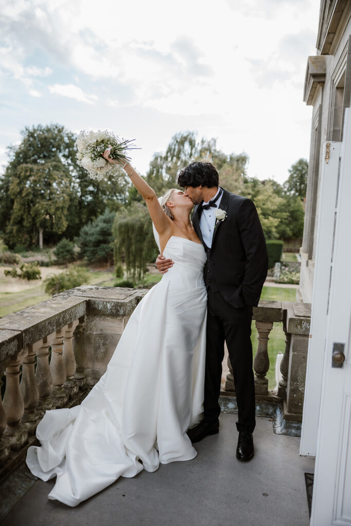 A bride in a white gown and a groom in a tuxedo kiss on a balcony. The bride holds a bouquet aloft, and gardens are visible in the background.