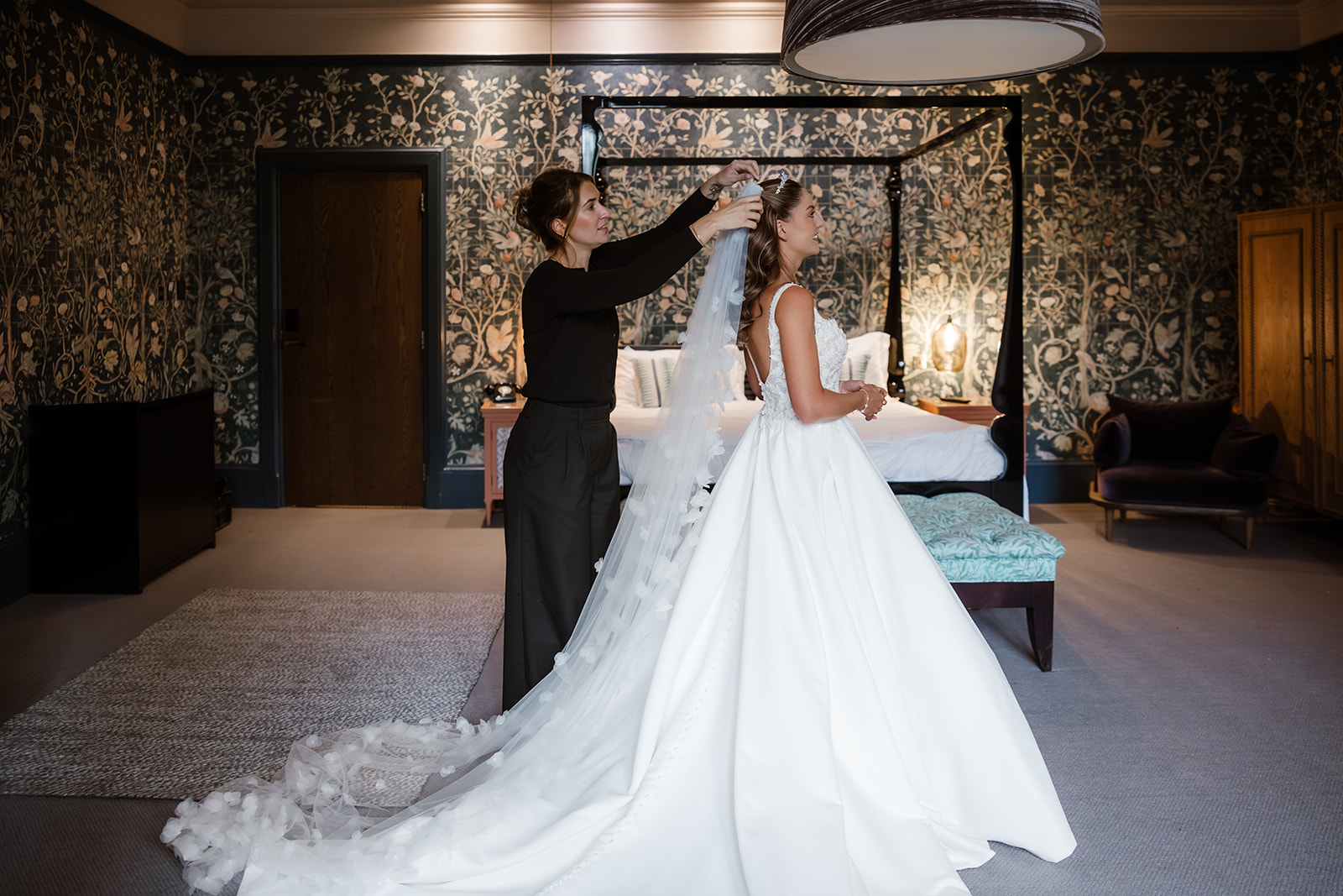 A woman adjusts the veil on a bride's head in a stylish, wallpapered bedroom with a four-poster bed.
