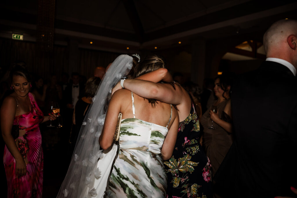 Two women, one in a wedding dress and veil and the other in a white and green dress, embrace on a crowded, dimly lit dance floor at an event.