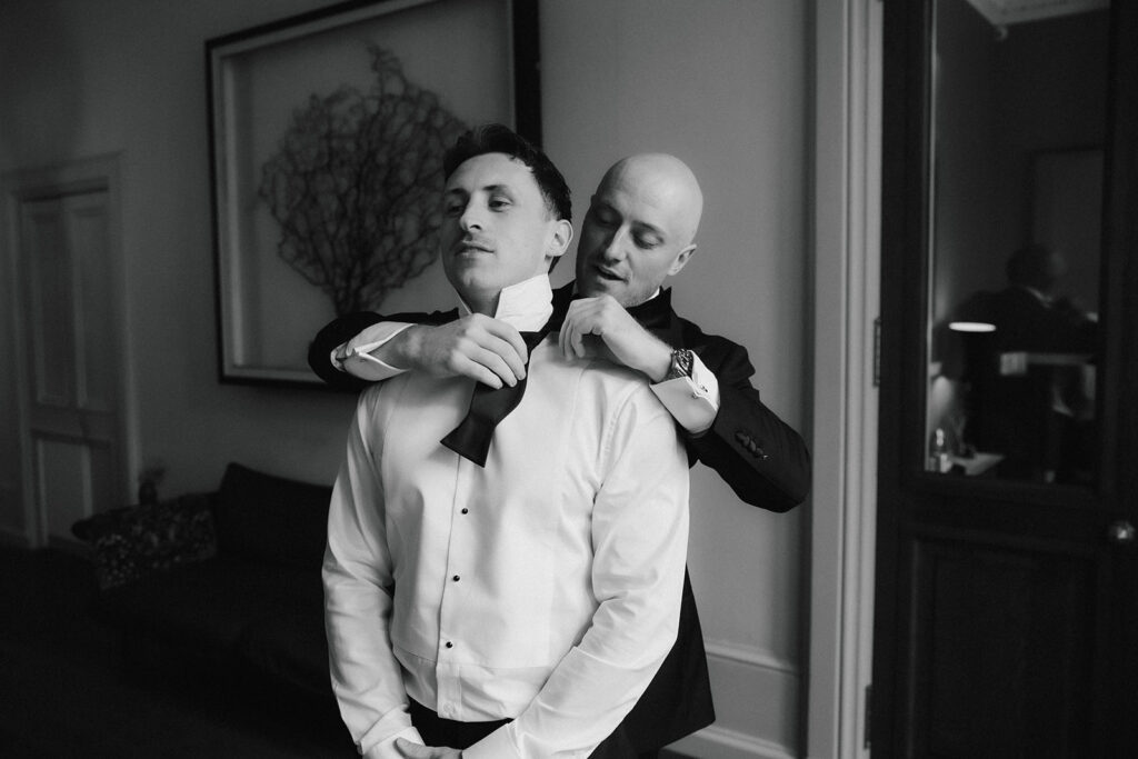 A man helps another man adjust his bow tie in a formal room, both dressed in suits.