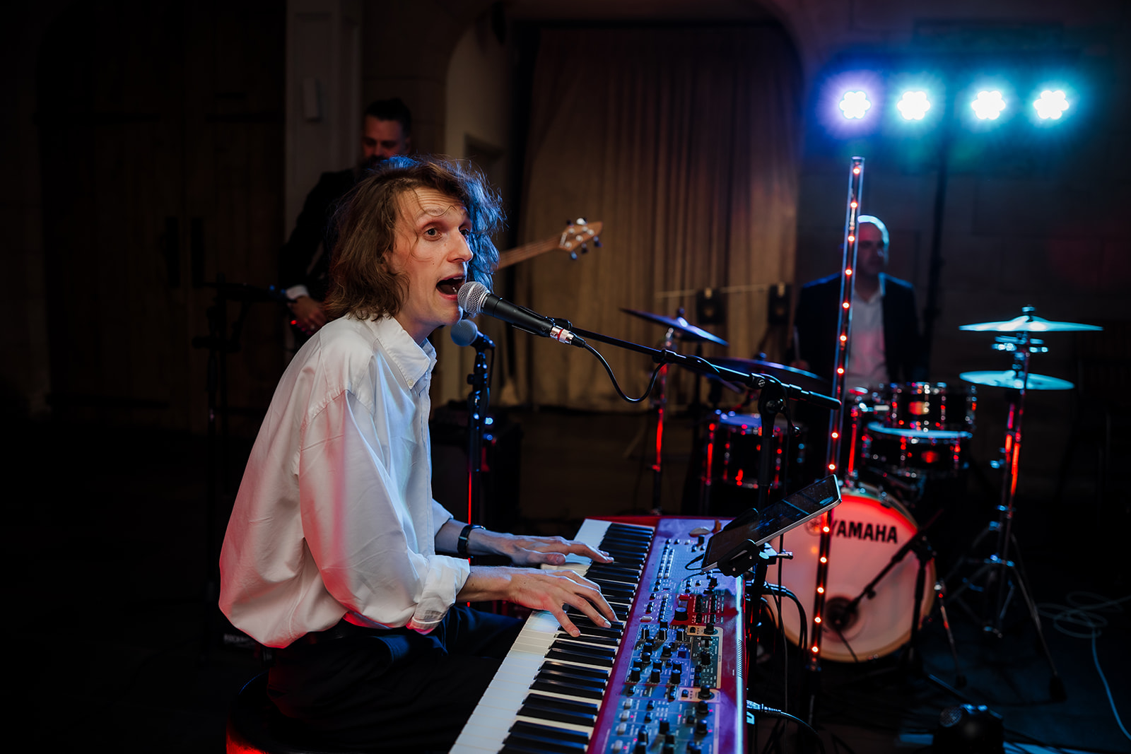 A musician in a white shirt plays a keyboard and sings into a microphone, with a drummer and bassist performing in the background under stage lighting.