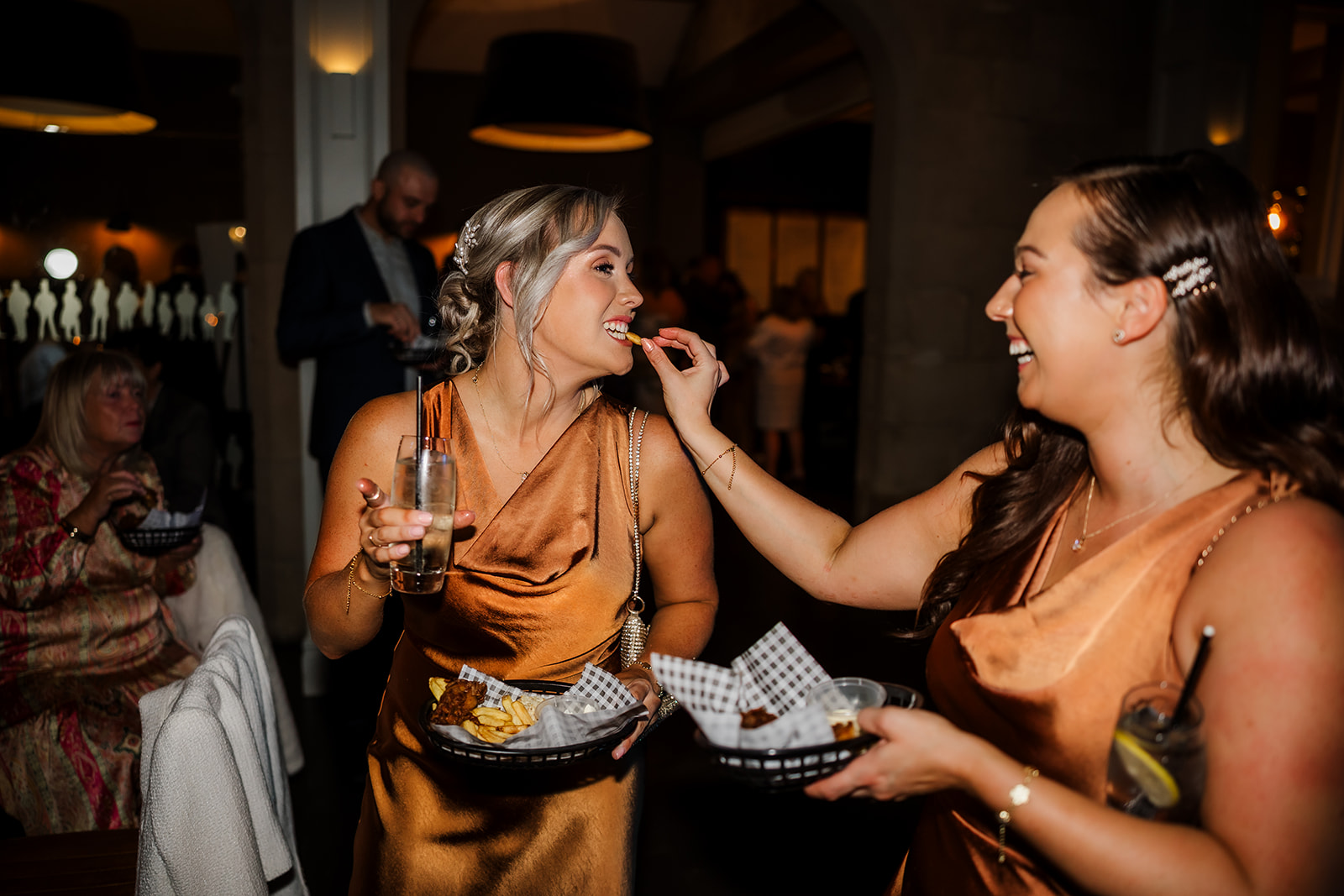 Two women in satin dresses smile as one feeds the other a chip at an indoor event; both hold food baskets and drinks, with guests visible in the background.