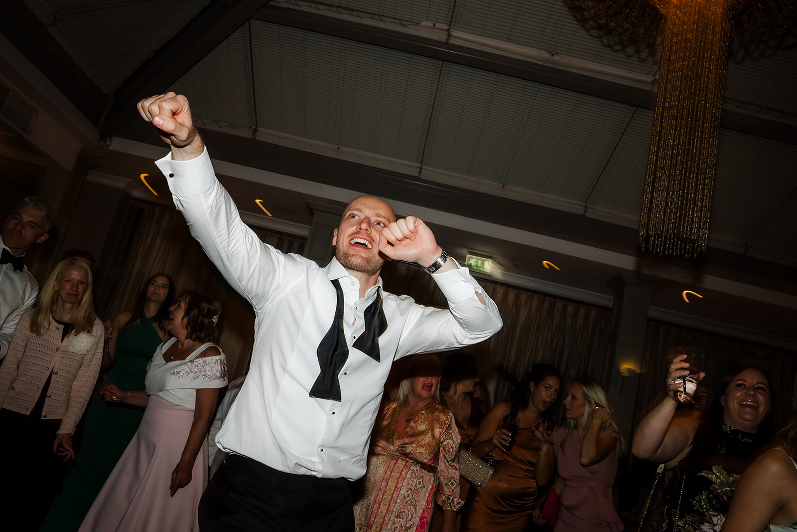 A man in a dress shirt and untied bow tie dances energetically at an indoor event, surrounded by a group of people in formal attire.
