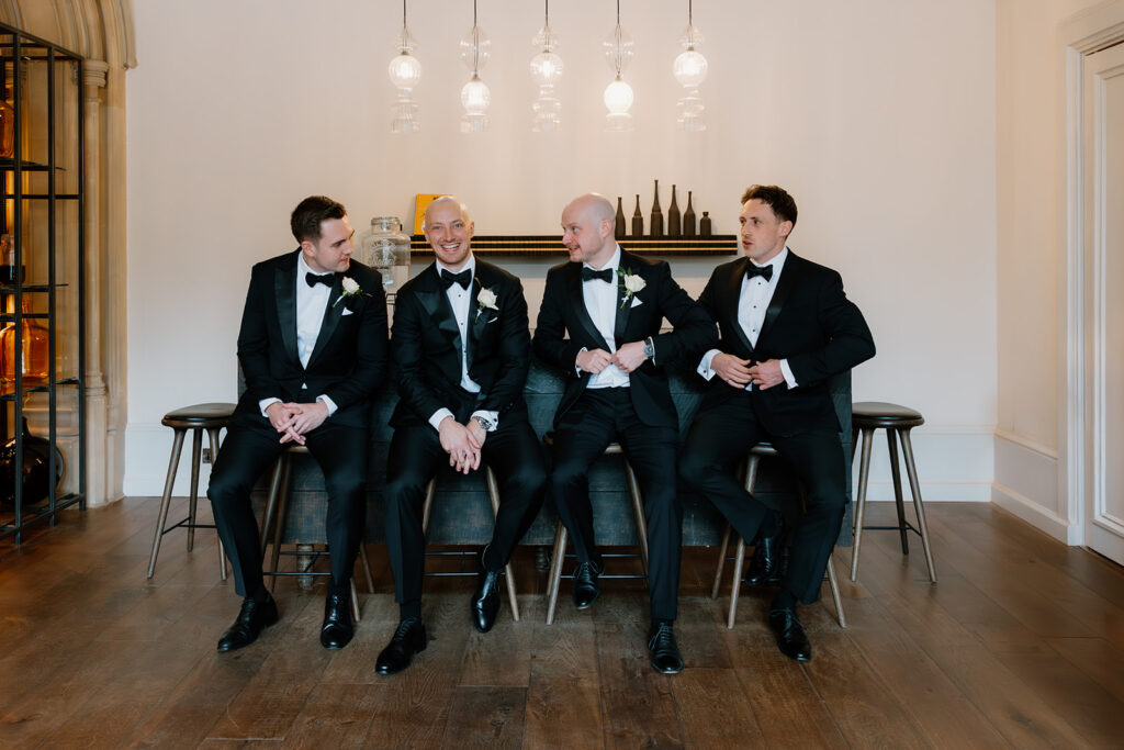 Four men, the groom and groomsmen, in black dinner suits sit on a sofa, smiling and talking in a modern room with wooden floors and pendant lights overhead.